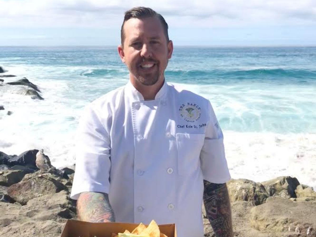 A person in a chef's coat is standing on a beach, holding a tray with tacos and chips, with waves crashing in the background.