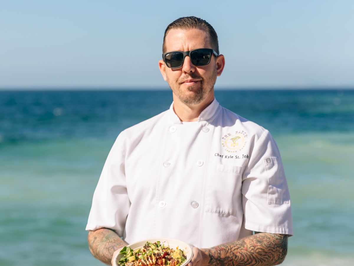A chef in a white jacket stands on a beach, holding a plate of food while the sea and clear sky form a scenic backdrop.