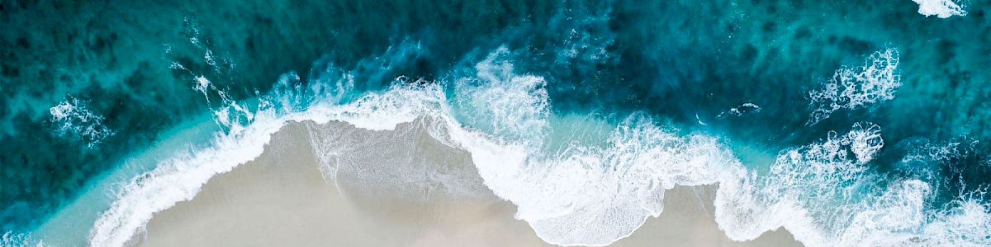 Aerial view of ocean waves crashing onto a sandy beach, showcasing various shades of blue and white from the water and foam, in a tranquil scene.