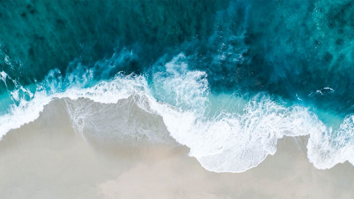 The image shows an aerial view of a beach with waves crashing onto the sandy shore, creating a contrast between the blue water and the light sand.