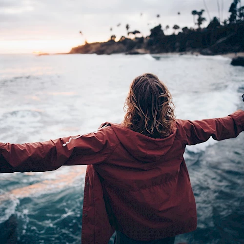 A person in a red jacket stands with arms outstretched, facing the ocean during sunset, holding a camera in one hand.