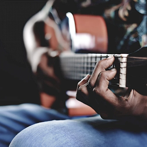 The image shows a person playing an acoustic guitar, focusing on their hands and the guitar strings, with a blurred background.