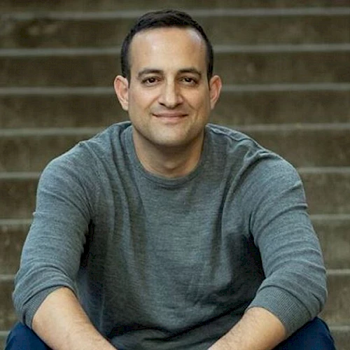 A person is sitting on steps, looking at the camera and smiling. They are wearing a gray long-sleeve shirt.