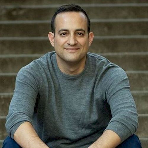 A man wearing a grey sweater is sitting on concrete steps, smiling at the camera, with his arms resting on his knees.