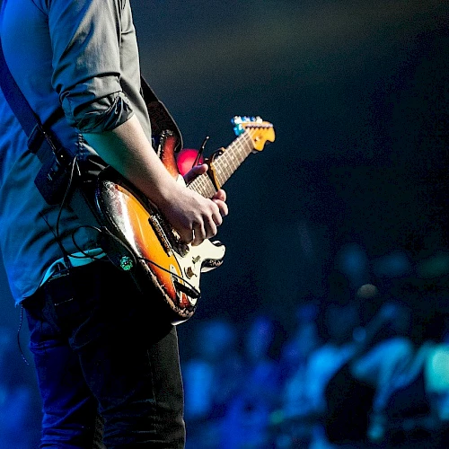 A person playing an electric guitar on stage, with a crowd of people watching in the background. The scene is lit with blue stage lights.