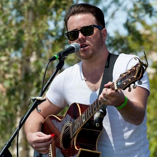A man wearing sunglasses and a white T-shirt plays an acoustic guitar and sings into a microphone outdoors, with trees in the background.