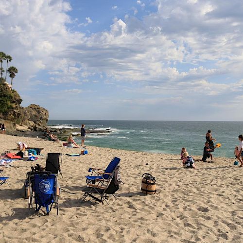 A beach scene with people relaxing, sitting on chairs, and walking on the sand. The sky is partly cloudy and the ocean is visible.