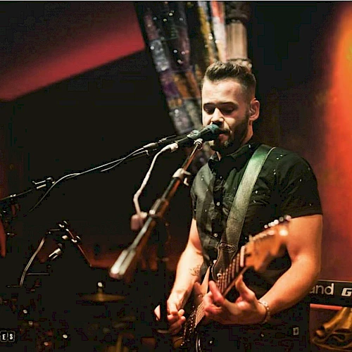 A musician is playing an electric guitar and singing into a microphone on stage, with ambient lighting and a colorful backdrop.