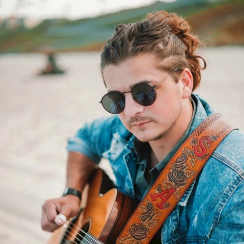 A person with sunglasses and a denim jacket is playing the guitar outdoors near a body of water, with blurred background.