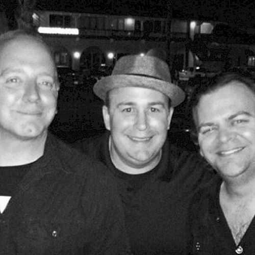 The image shows three smiling men standing closely together; one is wearing a hat. It appears to be night, with buildings visible in the background.