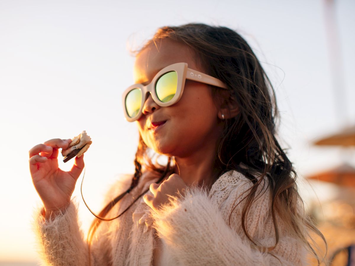 A young girl wearing large sunglasses holds a s'more while smiling, possibly enjoying a day at the beach with a soft-focus background.